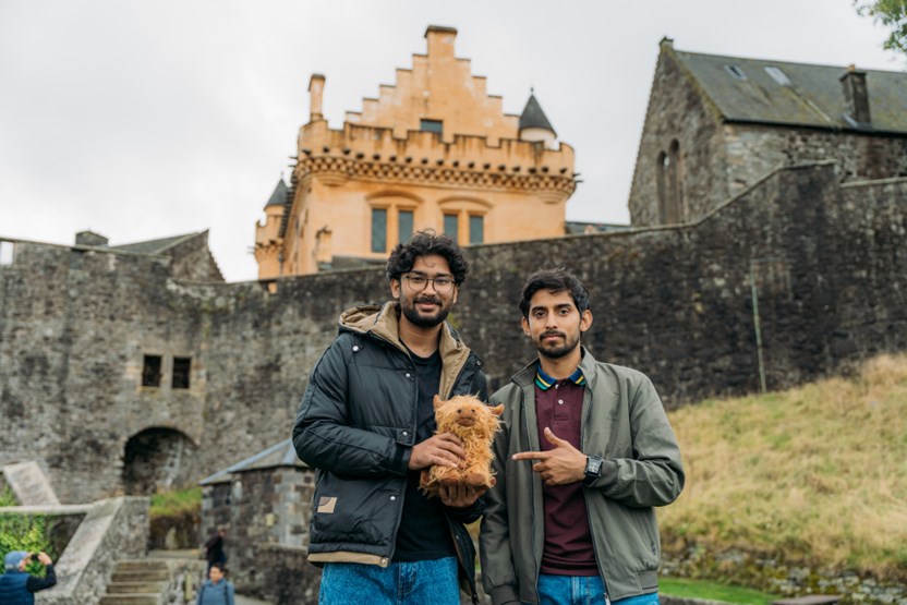 Two men holding a highland cow teddy bear, stand in front of a castle