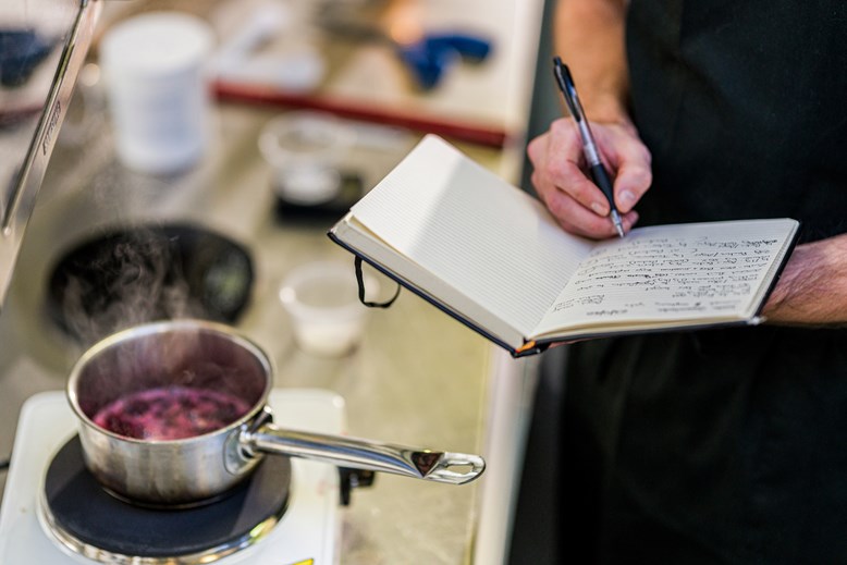 A person making notes in a notebook, while a pot simmers on a stove, in a kitchen.