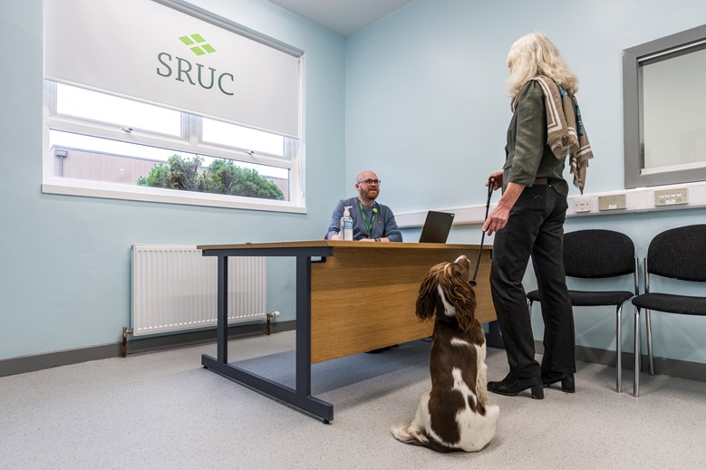 The reception room of a vet clinic. A man sitting at a desk with a computer, speaking to a customer with a dog on a leash.